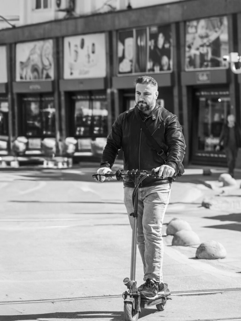 Man in a leather jacket riding an electric scooter on a city street, black and white